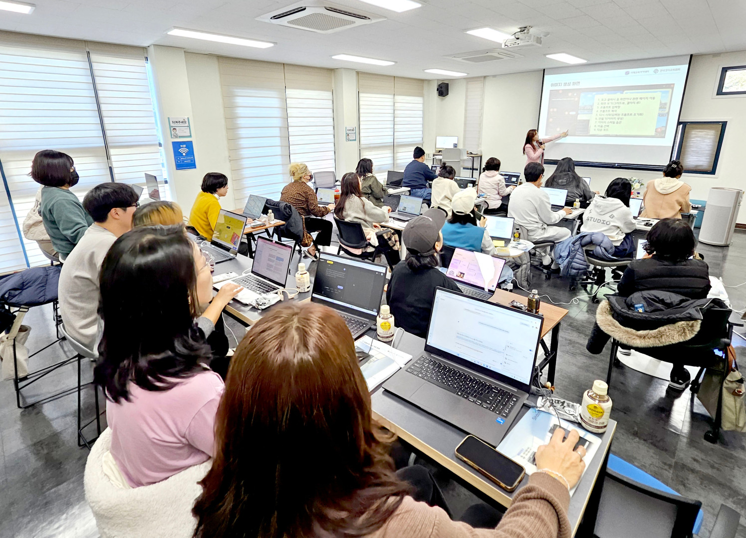 포항대학교 교육 현장에서 강사가 '이미지 생성 화면'에 대해 설명하는 모습. 전면 스크린에는 생성형 AI의 인터페이스와 함께 '1. 보고 클릭 시 각 화면이나 관련 페이지 이동', '2. 확인 및 체크(프롬프트, 갤러리 등)'와 같은 단계별 설명이 적혀 있습니다. 분홍색 상의를 입은 강사가 레이저 포인터로 스크린을 가리키며 강의하고 있으며, 교육생들은 개인 노트북을 통해 해당 내용을 실시간으로 확인하며 수업에 참여하고 있습니다.