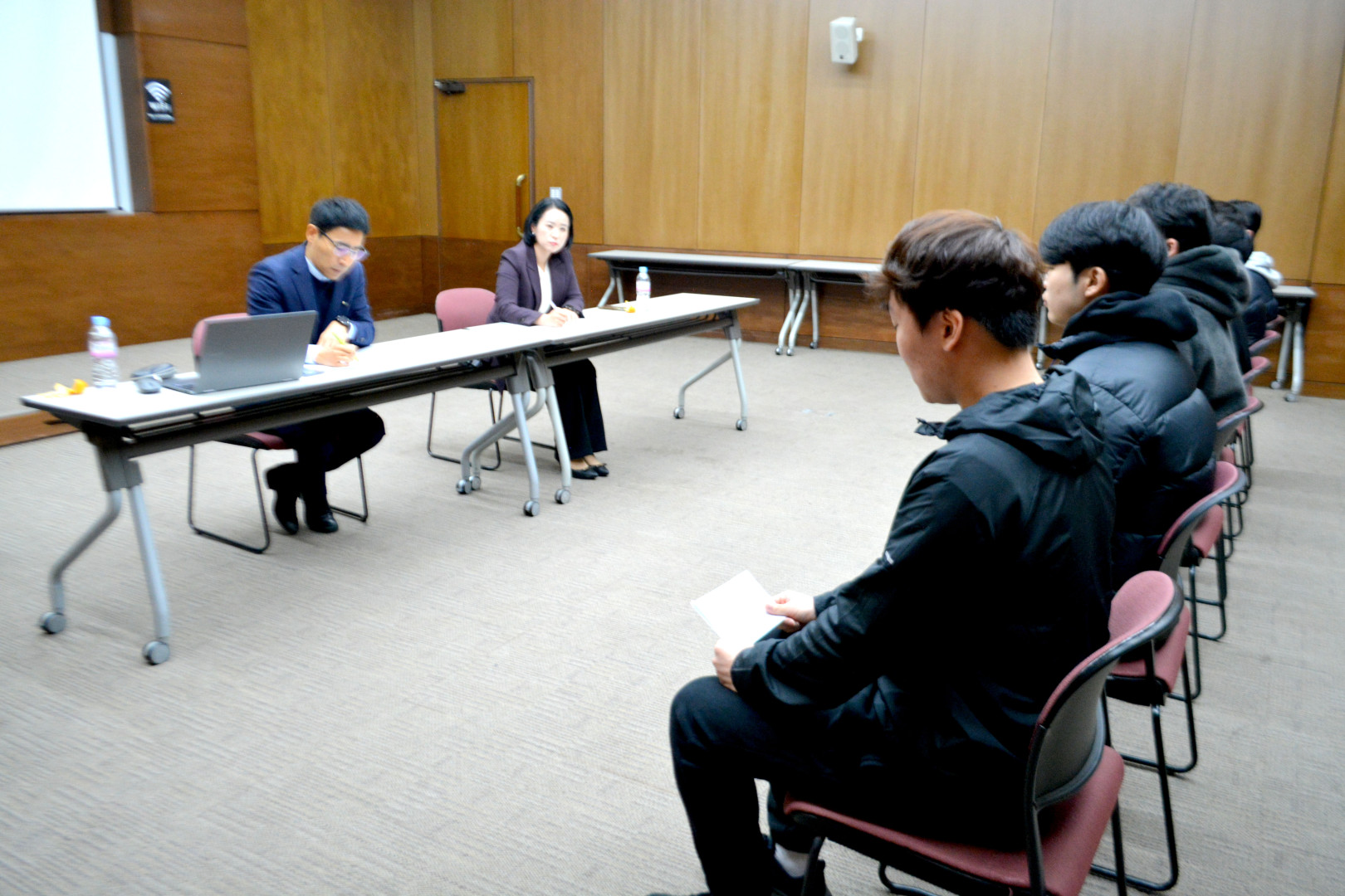 포항대학교 지역특화산업 취업캠프 모의 면접 진행 모습. 세미나실 내부에서 면접관 2명(남성 1명, 여성 1명)이 노트북과 서류가 놓인 긴 테이블에 앉아 있으며, 그 맞은편에 5명의 남성 학생들이 나란히 앉아 면접에 임하고 있습니다. 맨 앞쪽의 학생은 손에 면접 자료를 들고 대기하고 있으며, 전체적으로 진지하고 차분한 면접장 분위기가 느껴집니다.