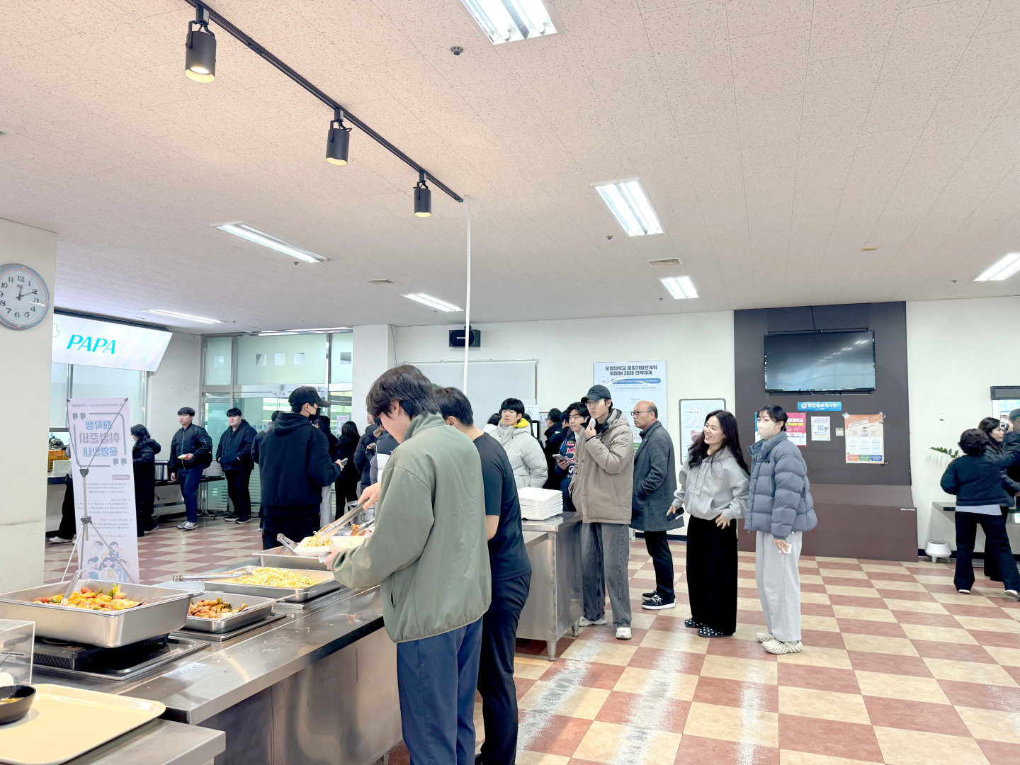 포항대학교 학생 식당 배식 현장 사진. 학생들이 자율 배식대 앞에서 줄을 서서 음식을 담고 있습니다. 배식대에는 탕수육, 볶음류 등 여러 가지 반찬이 담긴 대형 스테인리스 용기들이 놓여 있으며, 한 학생이 집게를 이용해 식판에 음식을 담고 있습니다. 뒤편으로는 식사를 위해 대기 중인 다른 학생들과 'PAPA'라고 적힌 간판이 보이며, 전체적으로 밝고 깨끗한 식당 내부 전경이 담겨 있습니다.
