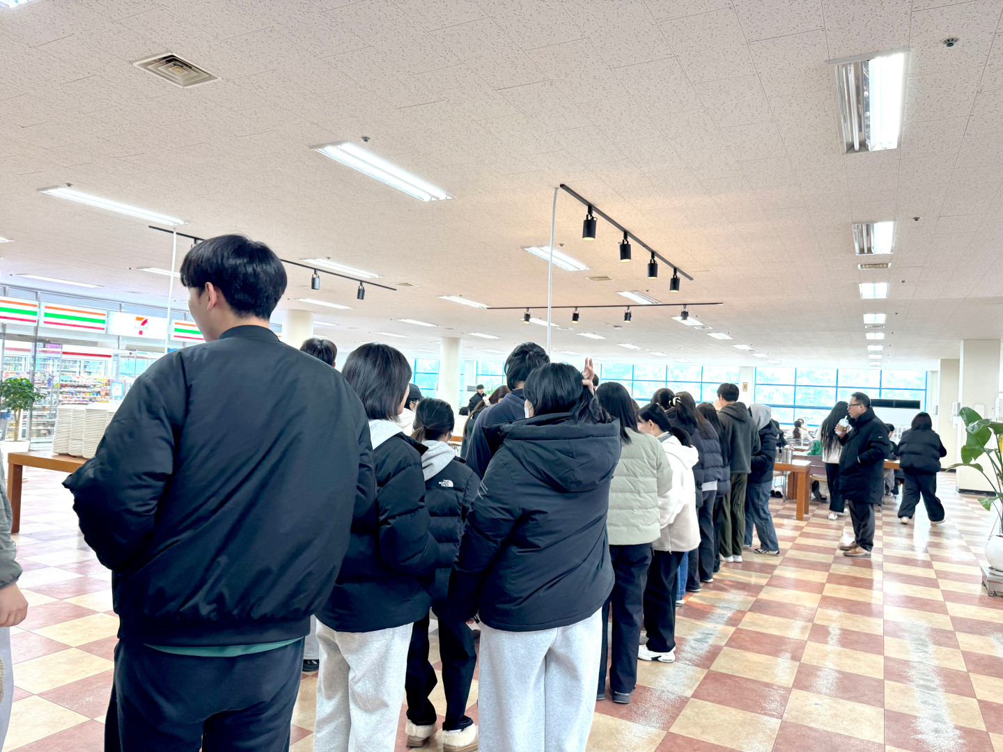 포항대학교 학생 식당 내부에서 학생들이 배식을 받기 위해 길게 줄을 서 있는 모습입니다. 검은색 패딩 등 겨울 옷차림을 한 학생들이 질서 정연하게 한 줄로 서 있으며, 화면 왼쪽 끝에는 세븐일레븐 편의점이 입점해 있는 모습이 보입니다. 넓은 식당 홀에는 사각형 테이블들이 배치되어 있고, 천장에는 일정한 간격으로 조명이 설치되어 있습니다.