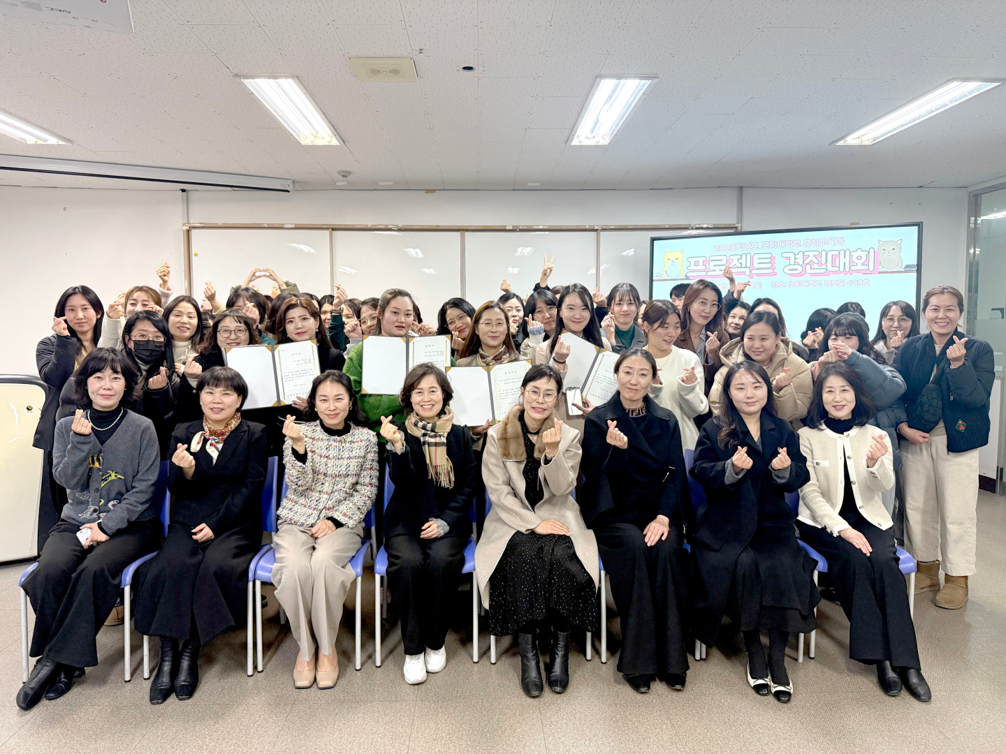 포항대학교 유아교육과 2025학년도 프로젝트 경진대회 단체 기념사진. 화면 오른쪽 상단 TV 스크린에는 '2025학년도 포항대학교 유아교육과 프로젝트 경진대회, 일시: 2025.11.28, 장소: 평보관 409호'라는 안내 문구가 띄워져 있습니다. 세미나실에서 교수진과 수십 명의 학생들이 모여 단체 사진을 찍고 있으며, 앞줄에 앉은 교수진과 뒷줄에 선 학생들 모두 손가락 하트 포즈를 취하며 웃고 있습니다. 중앙의 학생들은 상장으로 보이는 서류를 펼쳐 보이고 있습니다.