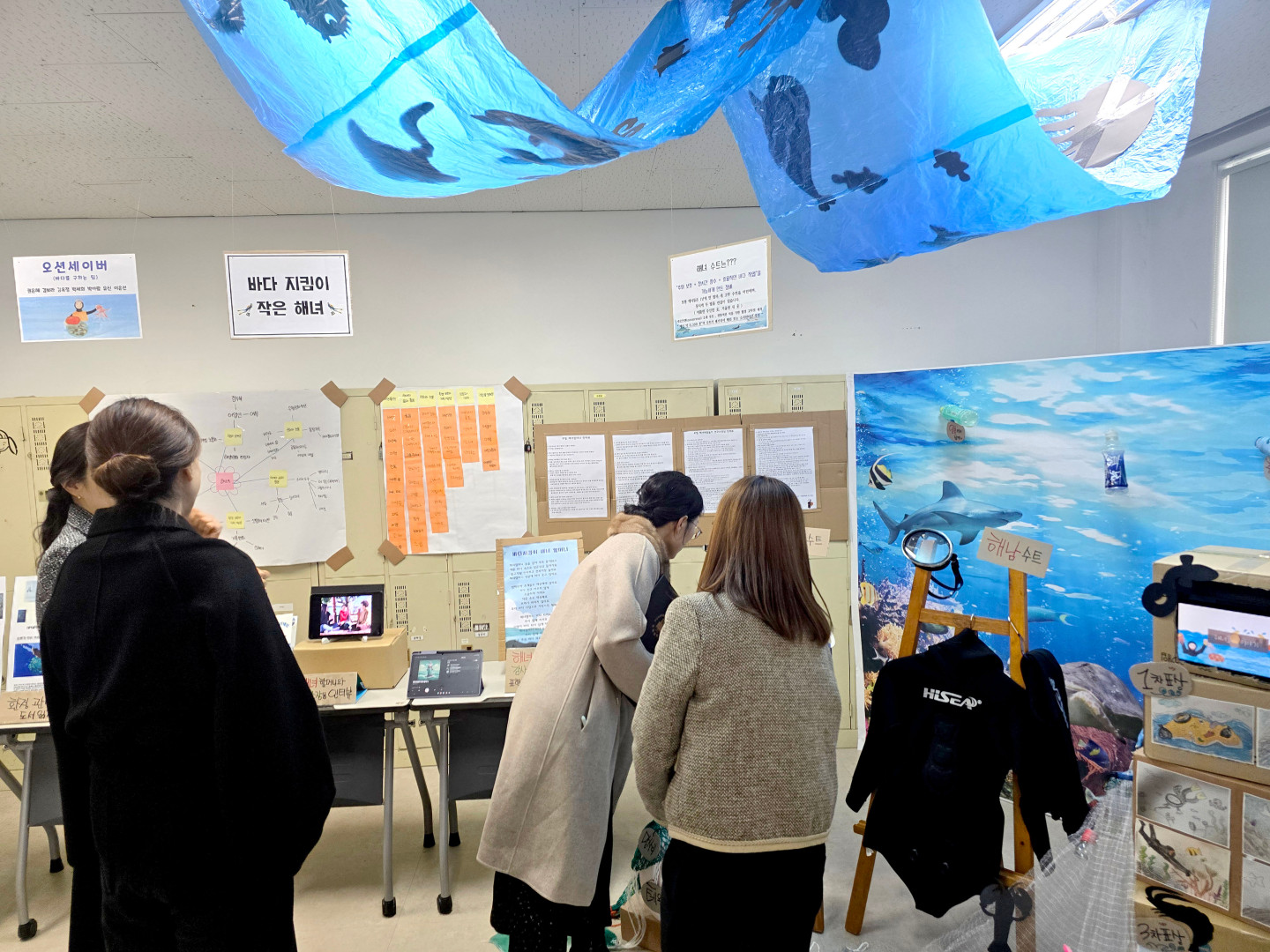 포항대학교 유아교육과 프로젝트 전시회 현장 사진. '오션세이버(바다를 구하는 팀)', '바다 지킴이 작은 해녀' 등의 팀별 프로젝트 결과물이 벽면과 테이블에 전시되어 있습니다. 천장에는 바다를 상징하는 파란색 비닐과 물고기 그림들이 매달려 있고, 벽면에는 마인드맵과 연구 자료들이 부착되어 있습니다. 전시 공간 한쪽에는 해녀 슈트와 물안경이 전시되어 있으며, 몇몇 학생들이 전시된 자료와 태블릿 PC 영상을 집중해서 관람하고 있습니다.