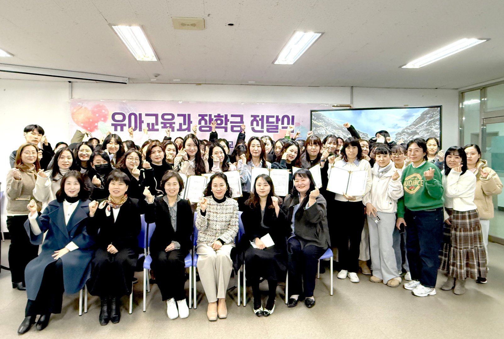 포항대학교 유아교육과 장학금 전달식 기념사진. 배경에는 '유아교육과 장학금 전달식'이라고 적힌 분홍색 현수막이 걸려 있습니다. 세미나실로 보이는 장소에서 수십 명의 학생들과 교수진이 모여 단체 사진을 촬영하고 있습니다. 앞줄에 앉은 6명의 여성 관계자들과 뒷줄에 서 있는 많은 학생이 손가락 하트 포즈를 취하거나 주먹을 쥐고 파이팅 포즈를 취하며 밝게 웃고 있습니다. 중앙의 몇몇 학생들은 수여받은 장학 증서를 펼쳐 보이고 있습니다.